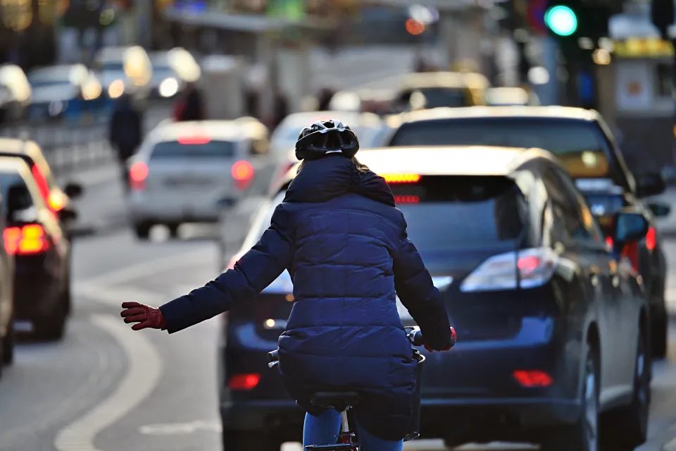 Rear view of woman in blue coat riding bike on busy road signals with left hand to merge into lane of traffic.